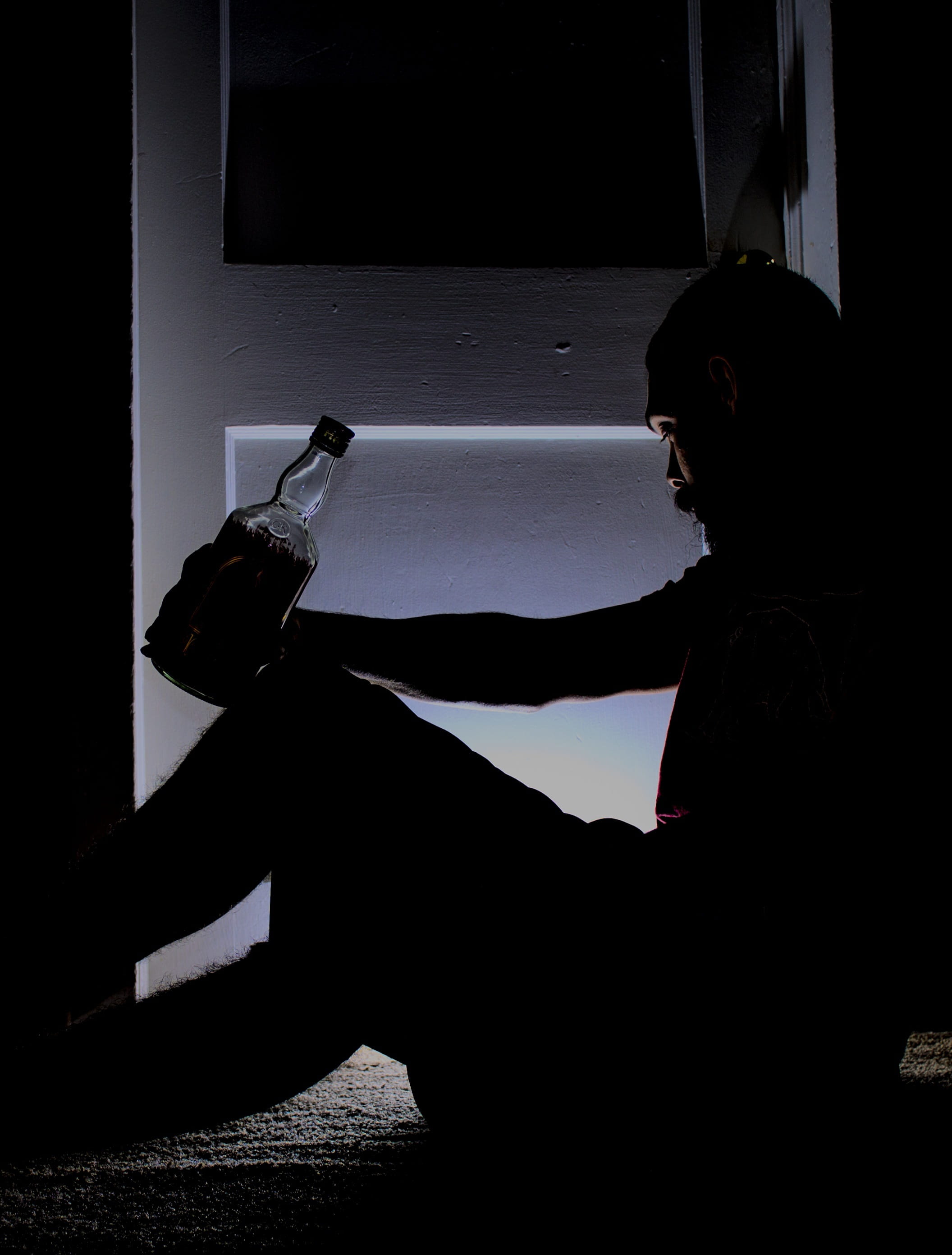 man sitting next to door holding bottle of liquor, drinking, lonely