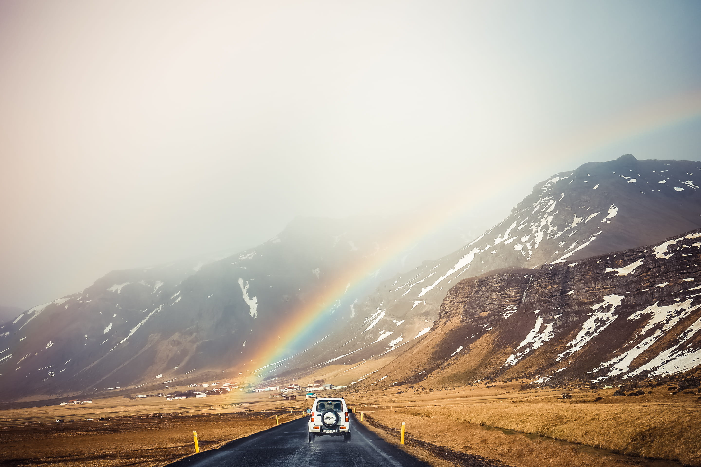 white car travelling near mountains with rainbow, white car traveling at city road in a distance of a mountain with rainbow