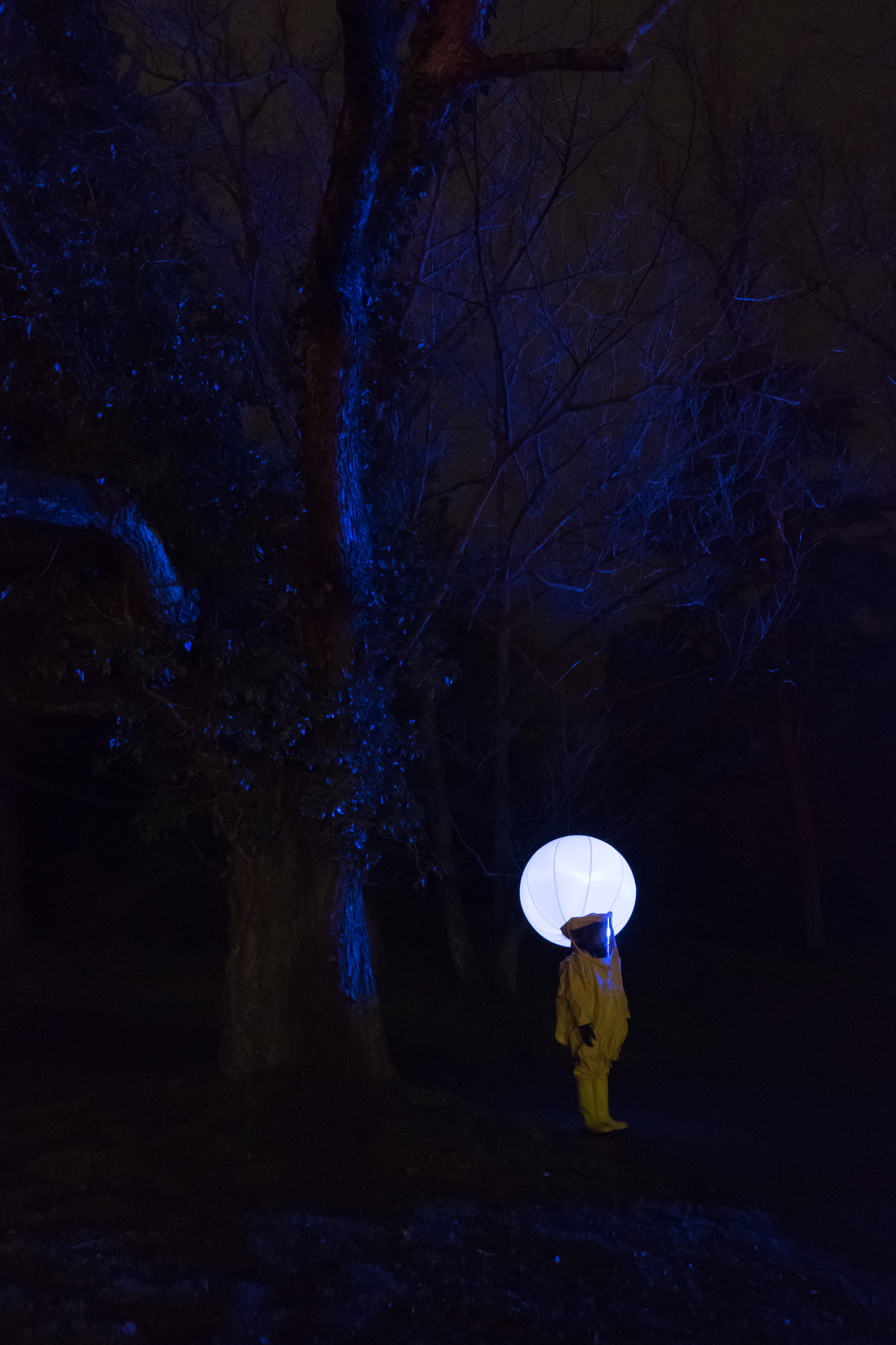 person in white outfit standing under leafless tree at nighttime, person wearing hazmat suit standing near tree