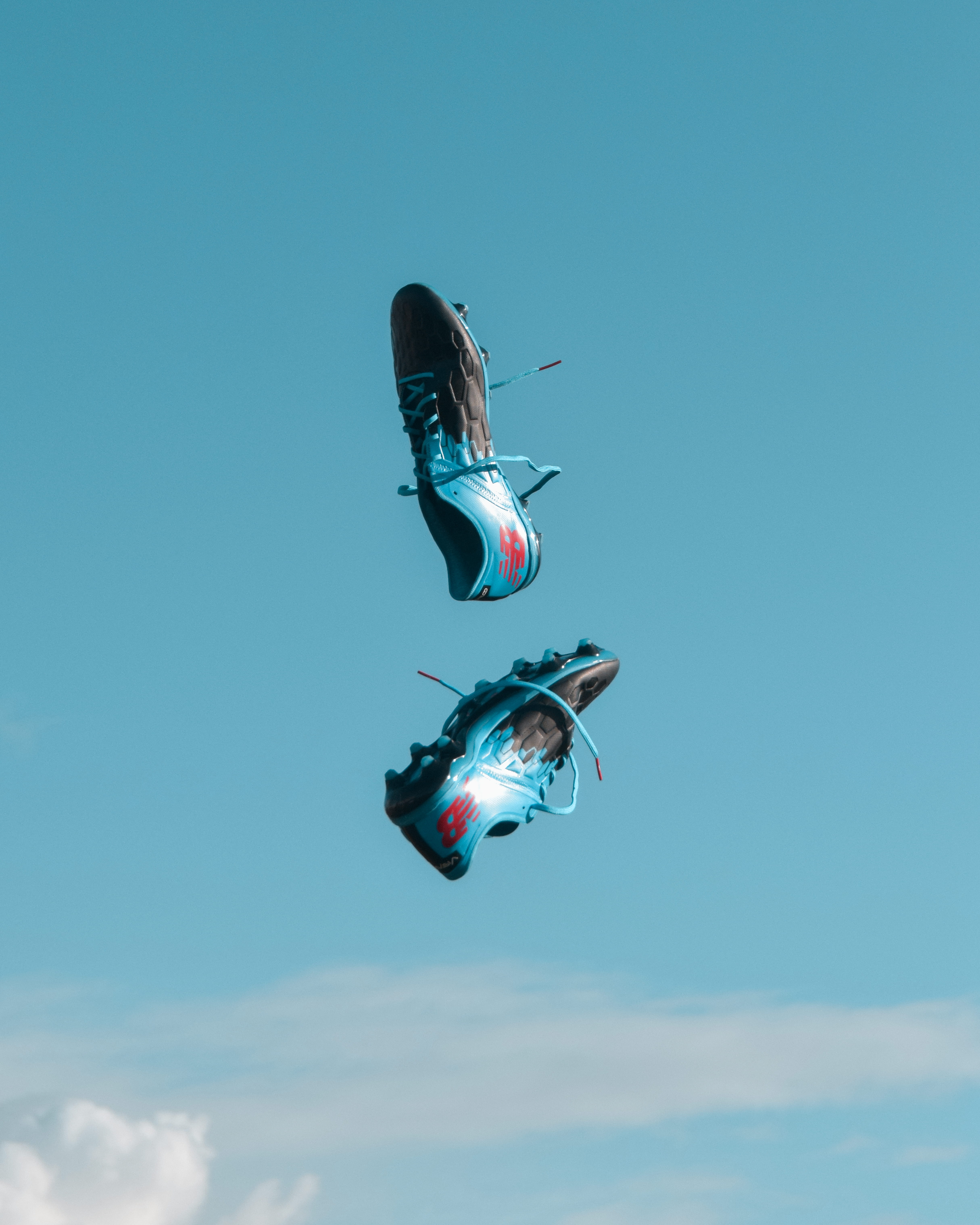 pair of blue-and-black New Balance cleats in the air under teal sky with white clouds, pair of black-and-blue New Balance cleats in air