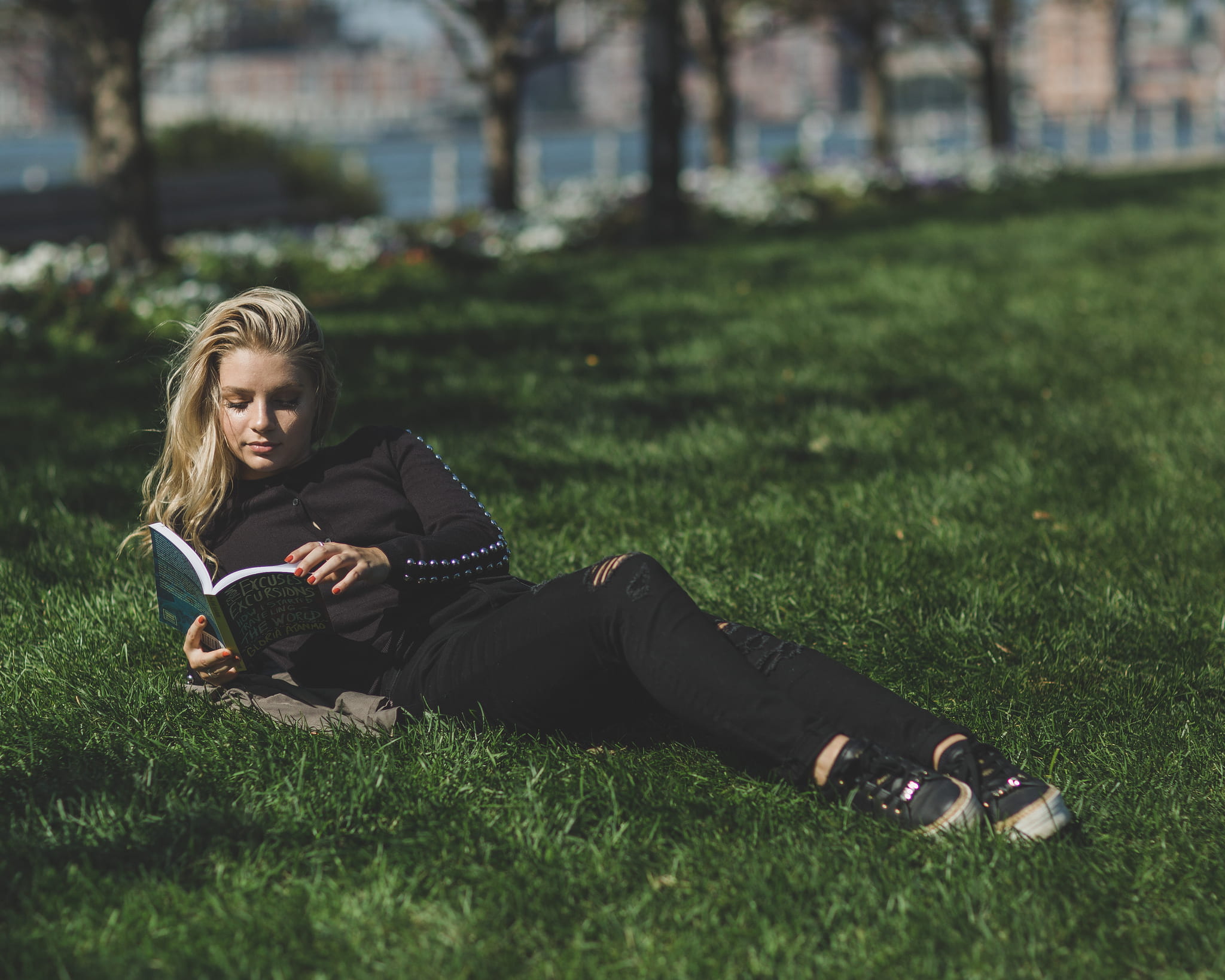 woman reading book leaning on green grass field, woman in black long-sleeved shirt and black pants lying on green grass while reading during daytime