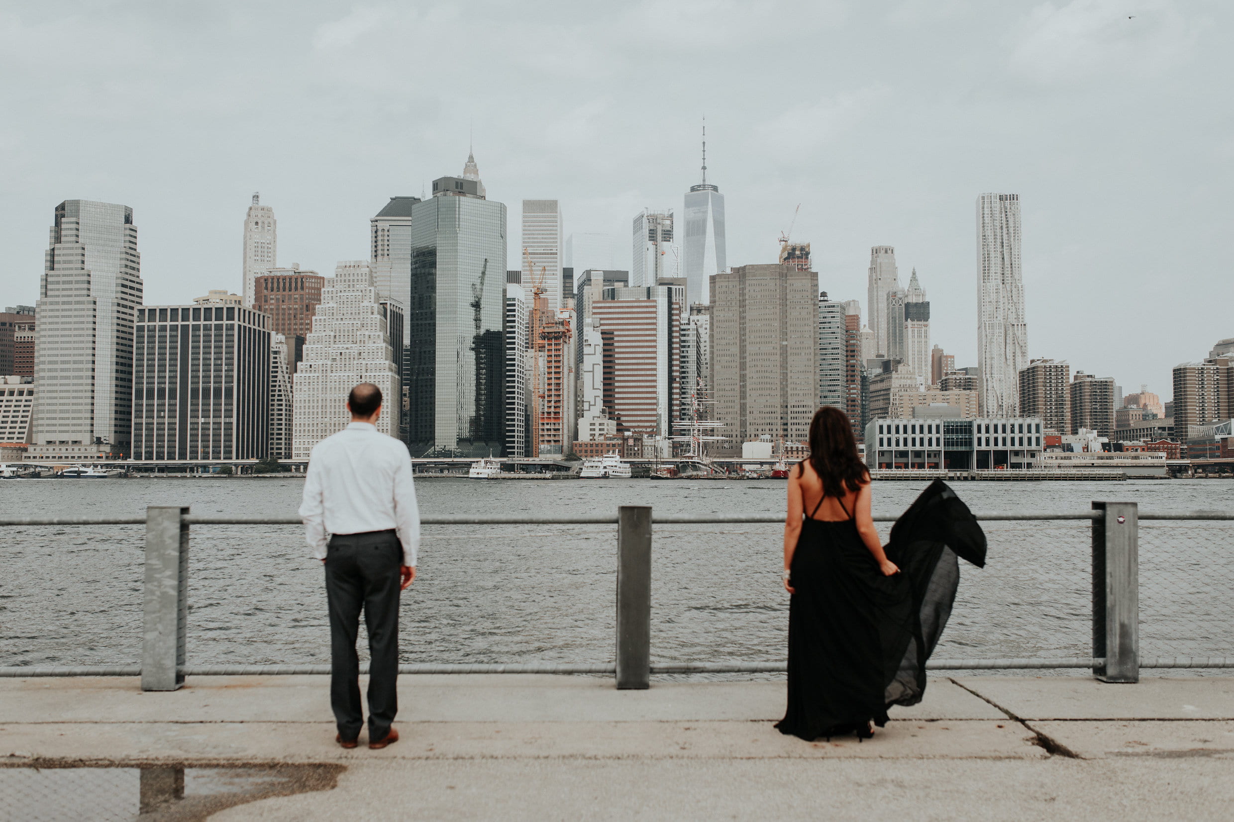 man and woman standing near body of water facing city buildings