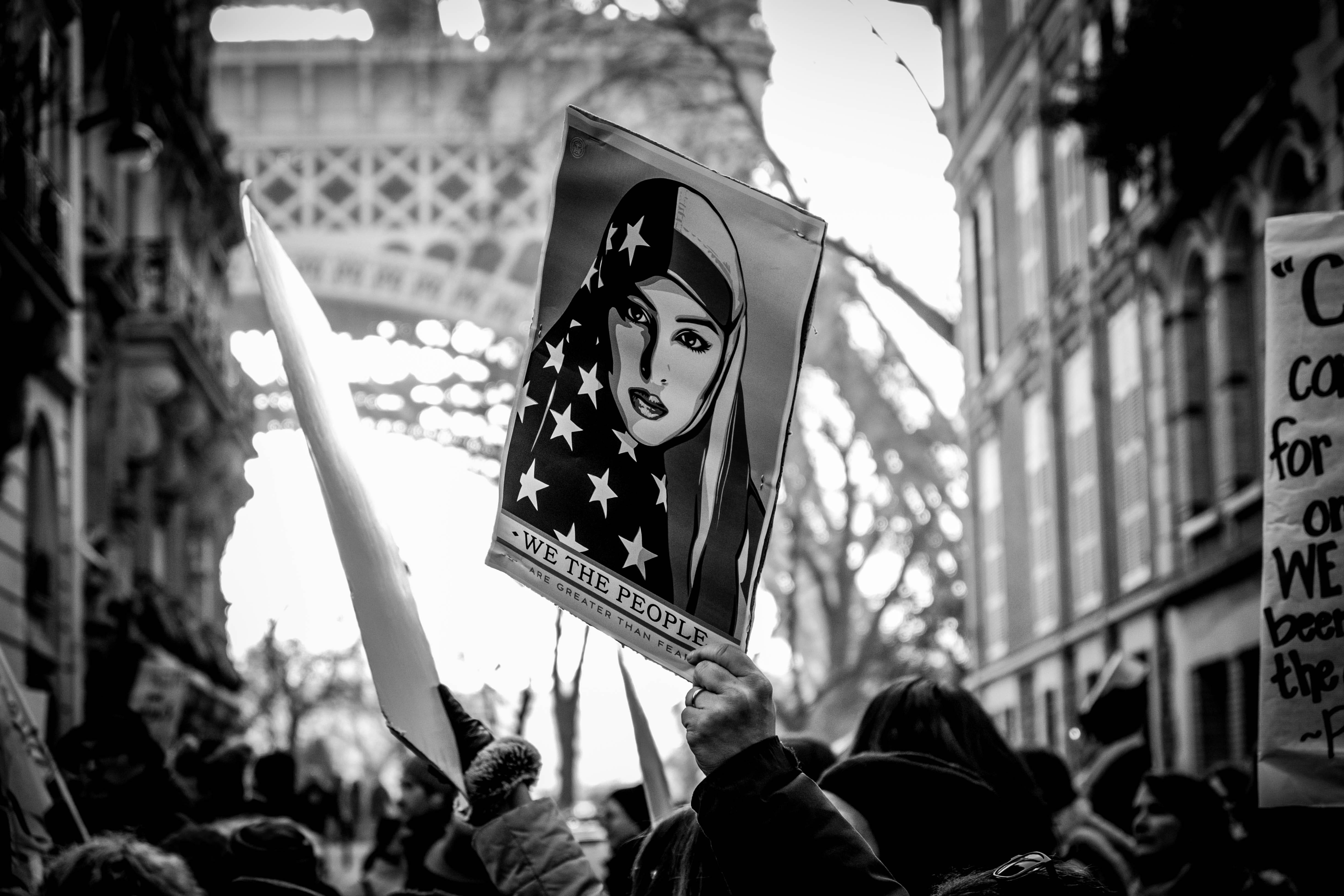 grayscale photography of woman face poster wave on air by people near Eiffel Tower, grayscale photography of person holding We The People banners