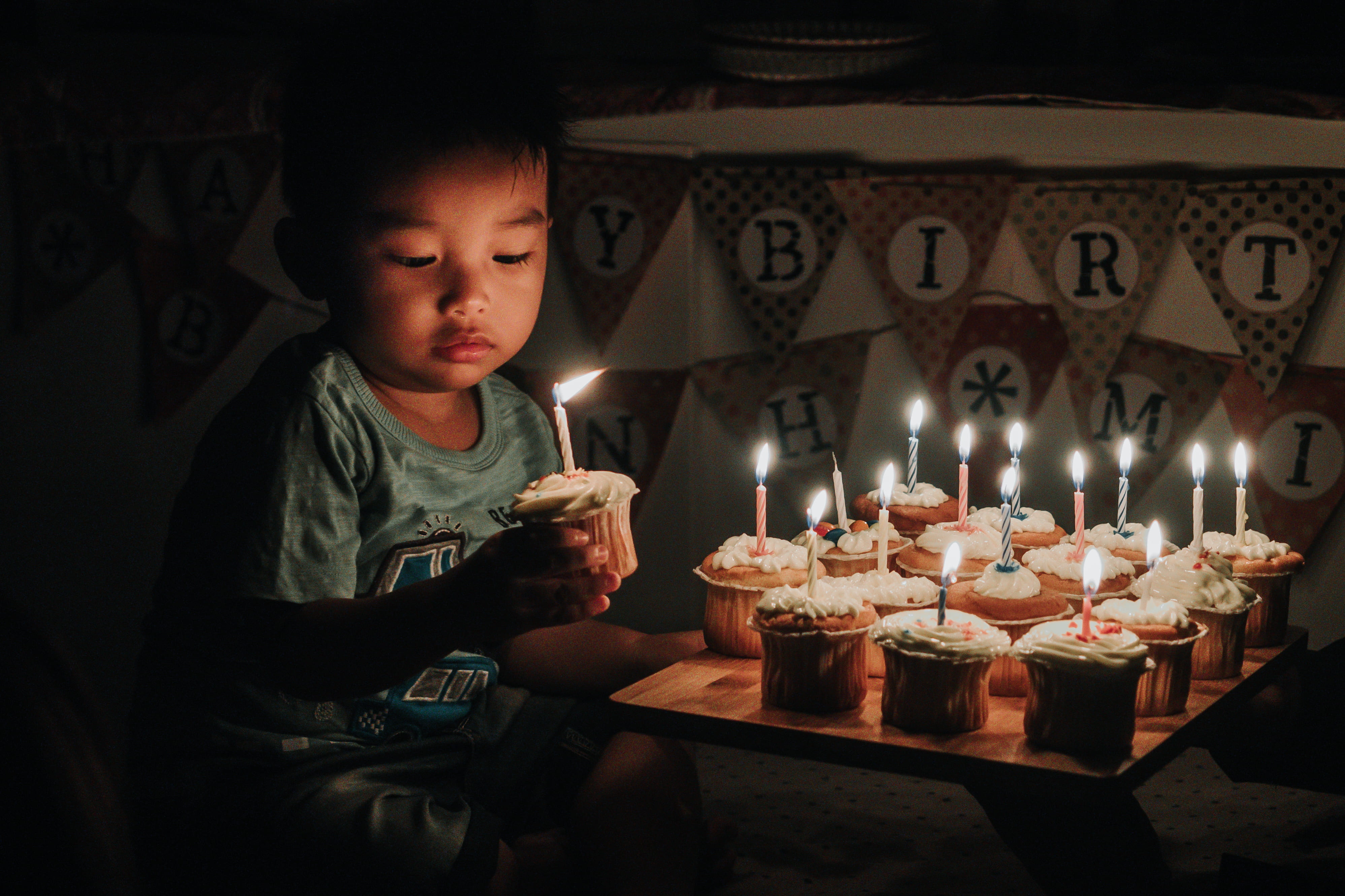 boy holding one of many cupcakes with lit candles, boy holding cupcake with lightning candle beside happy birthday buntings