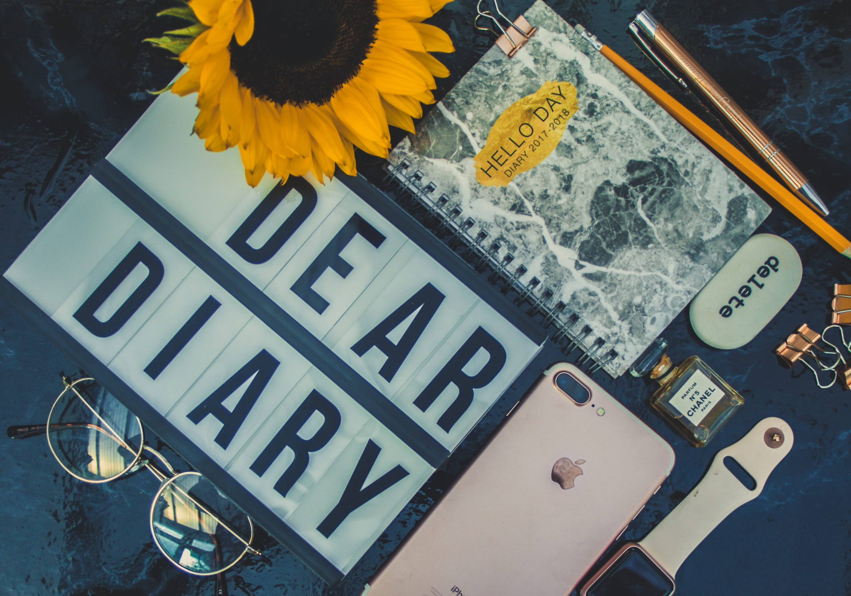 flay lay photography of a rose gold iPhone 7 Plus, Apple Watch, eyeglasses, notebook and sunflower