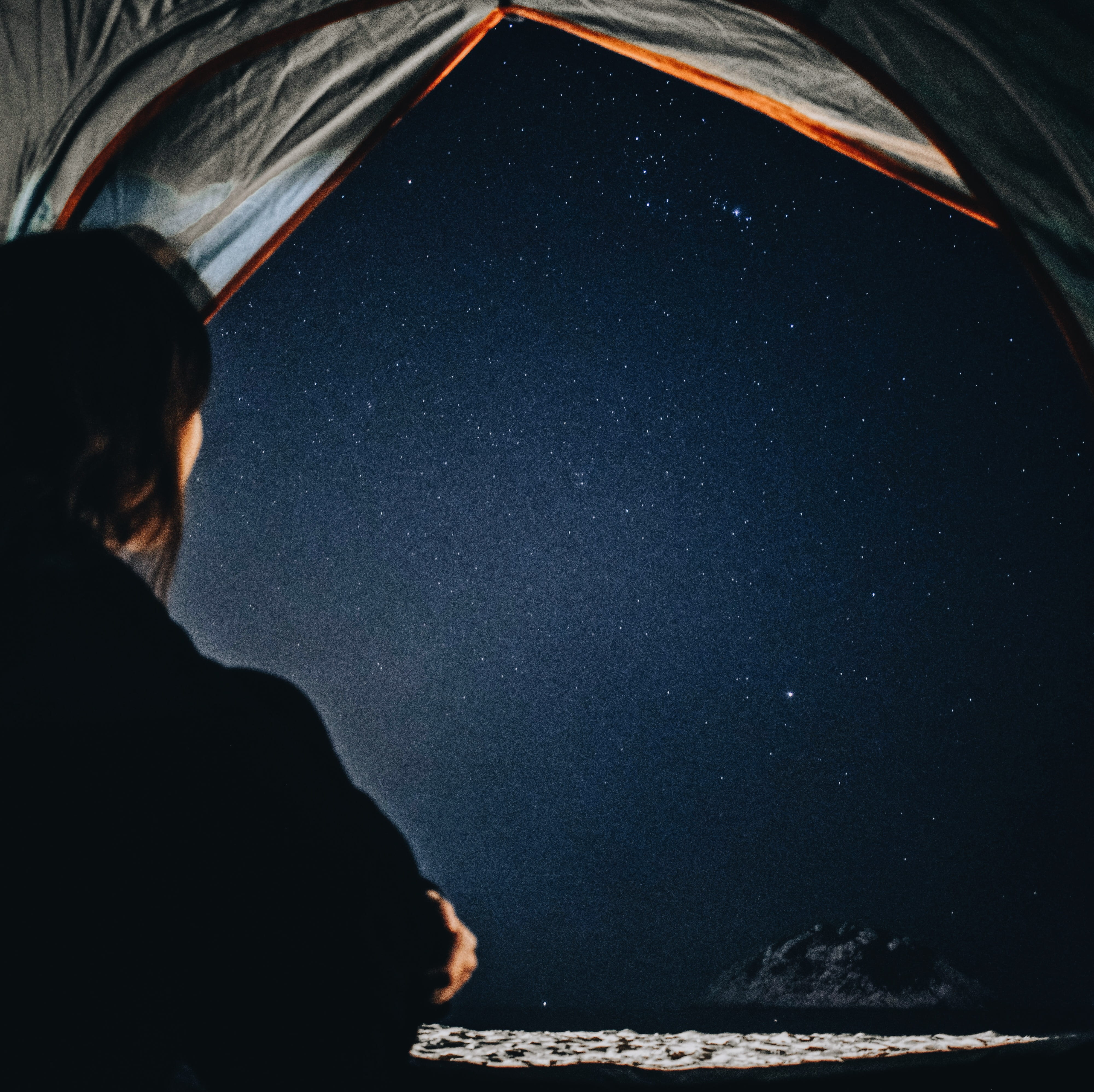 woman staring at stars at sky at night, person wearing black coat sitting inside gray camping tent with view of seashore during nighttime
