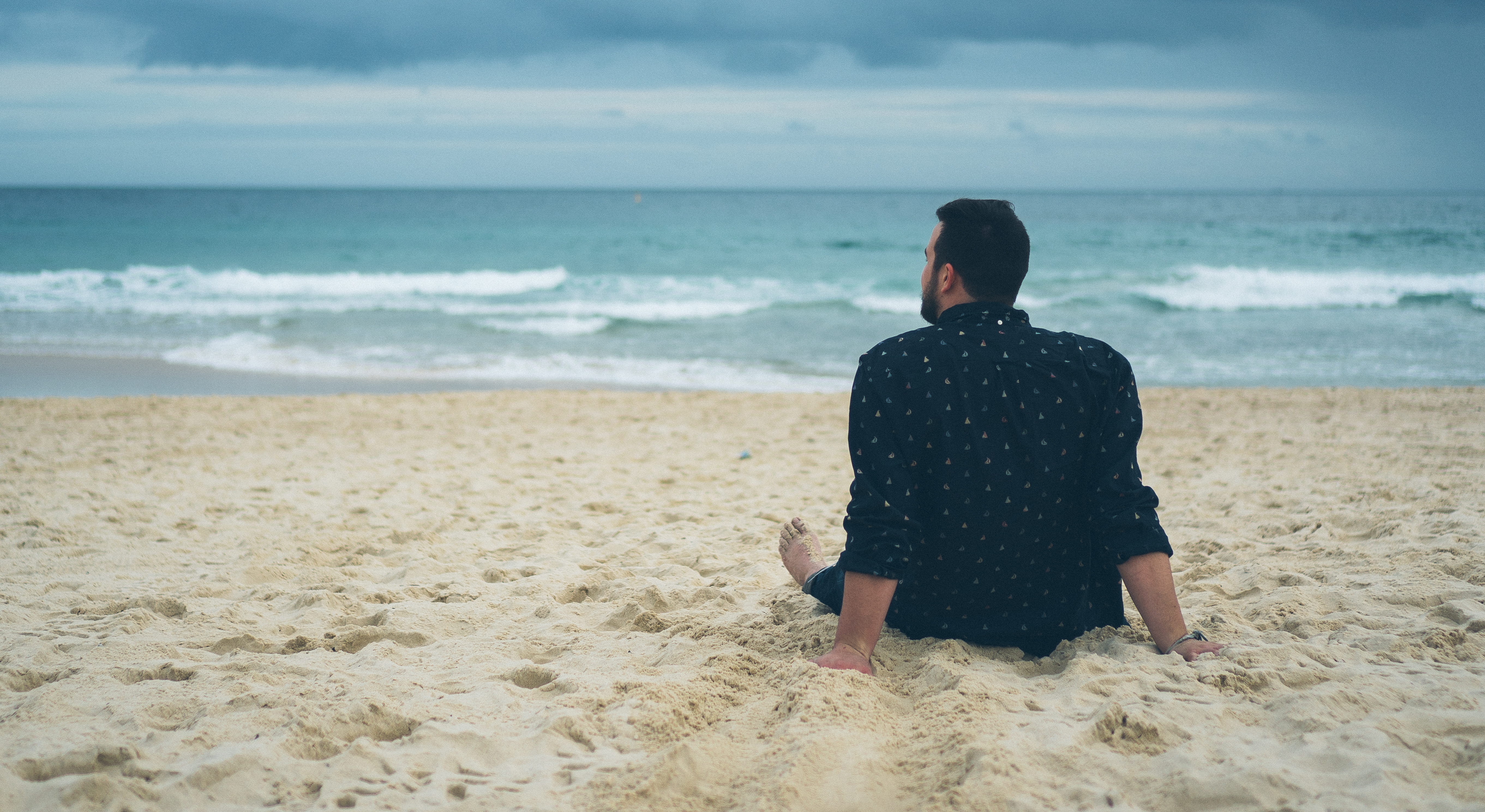 man sitting on beach sand, man sitting on seashore during cloudy sky