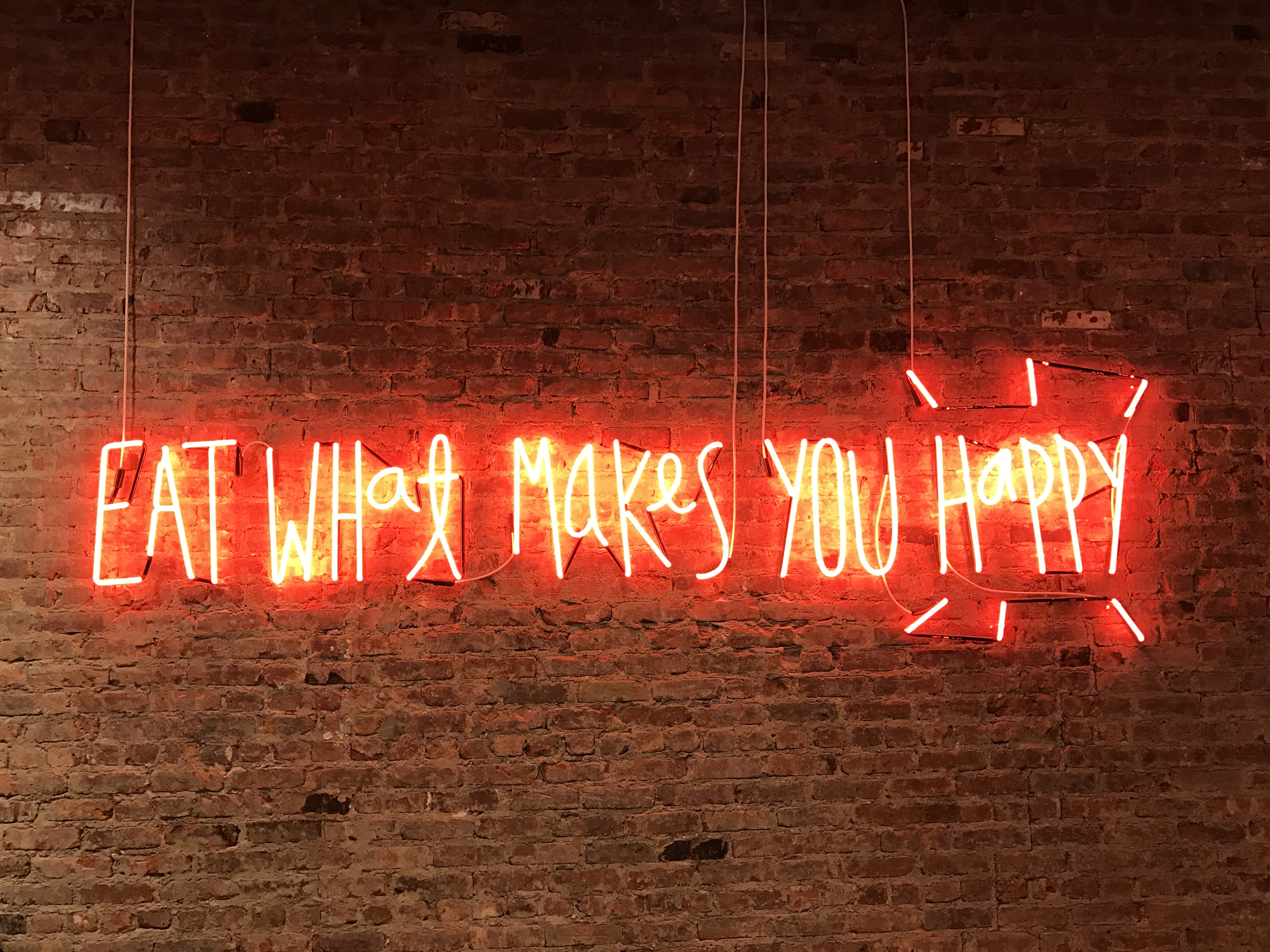 lighted eat what makes you happy neon signage, brown brick wall with quotes text overlay