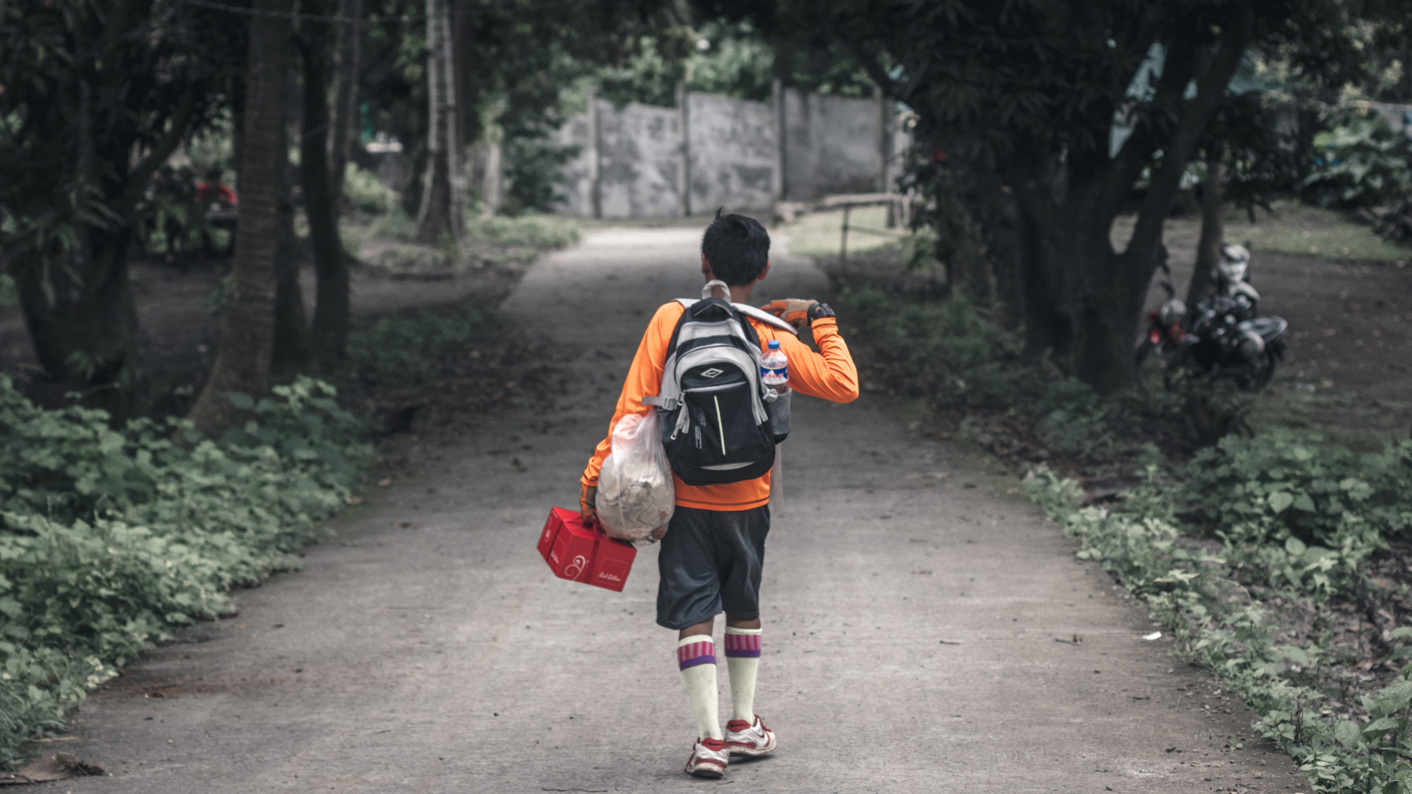 boy with backpack walking on pathway between trees, boy in orange long-sleeved shirt carrying backpack walking on pathway