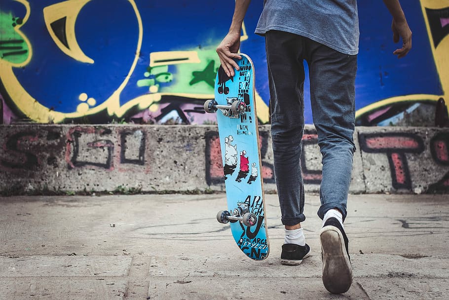 person holding blue skateboard walking near graffiti, man holding skateboard