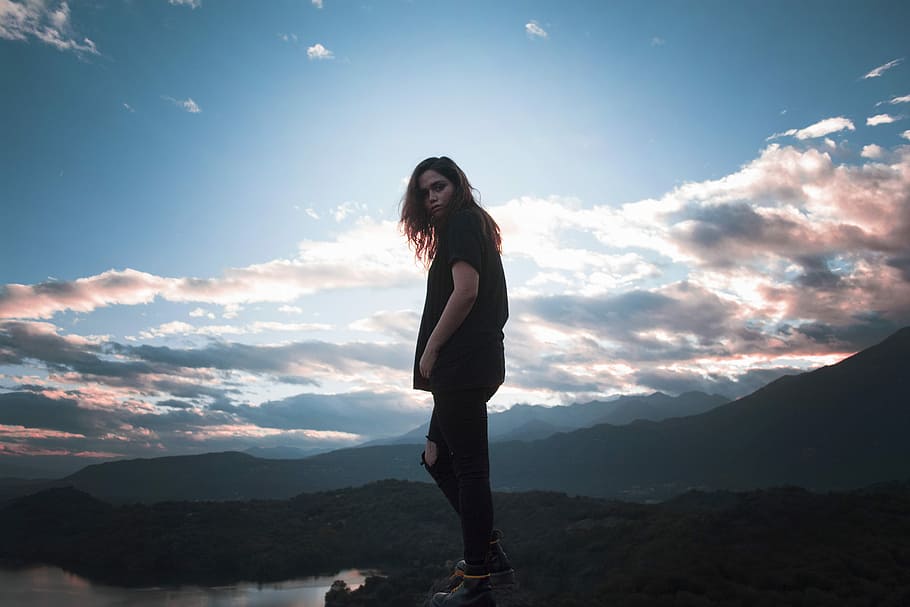 woman putting her hand in pocket standing near mountain, woman on cliff wearing black shirt HD wallpaper