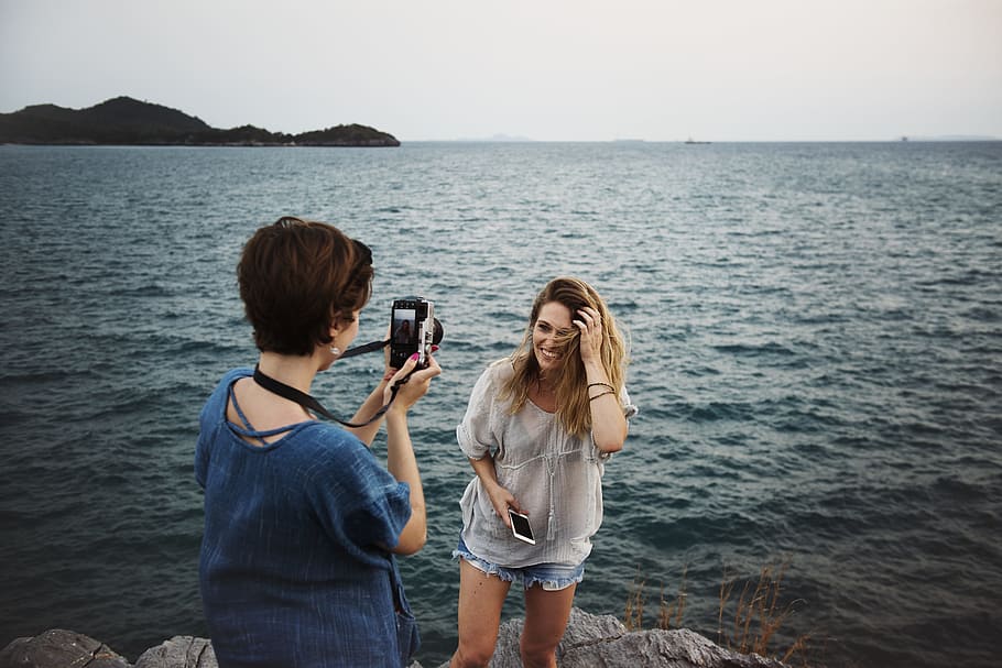 short haired woman in blue top taking photo of another woman in white top standing beside body of water, HD wallpaper