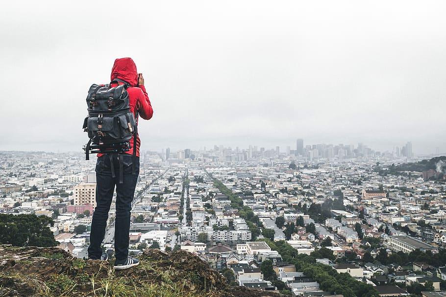 person standing on cliff over city, person in red hoodie and black pants with backpack standing at the top of mountain, HD wallpaper