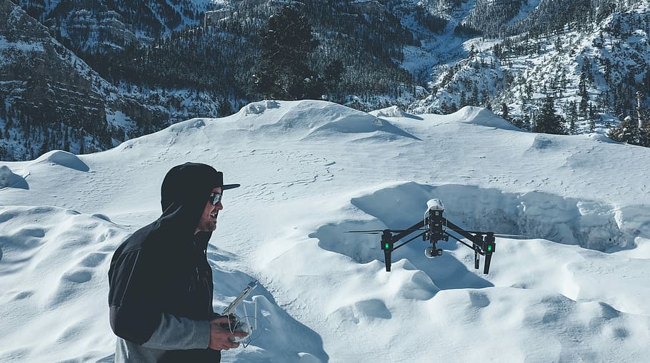 man wearing pull-over jacket infront of black quadcopter on snow field mountain HD wallpaper