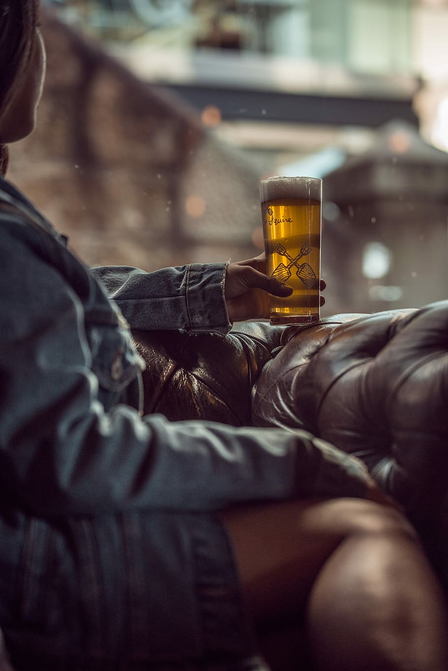 woman sitting on tufted brown leather chair holding pilsner glass filled with beer
