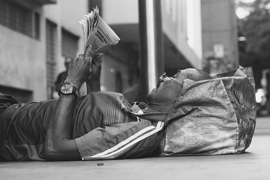 man lying on sidewalk reading newspaper, grayscale photo of man lying on floor while reading on newspaper