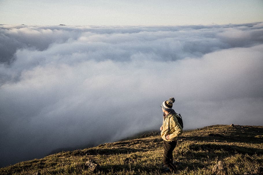 person inserting both hands inside jacket pockets while looking right side, man standing on cliff with clouds overlooking HD wallpaper