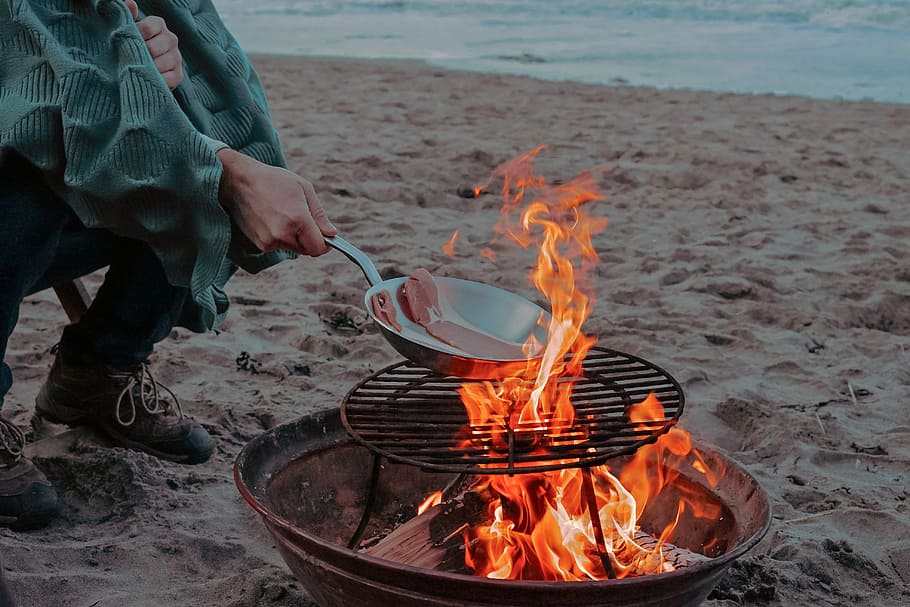 person cooking meat on bonfire near shoreline, person grilling meat near beach