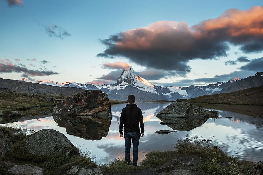 man standing on rocky cliff facing body of water, photo of man standing near body of water HD wallpaper