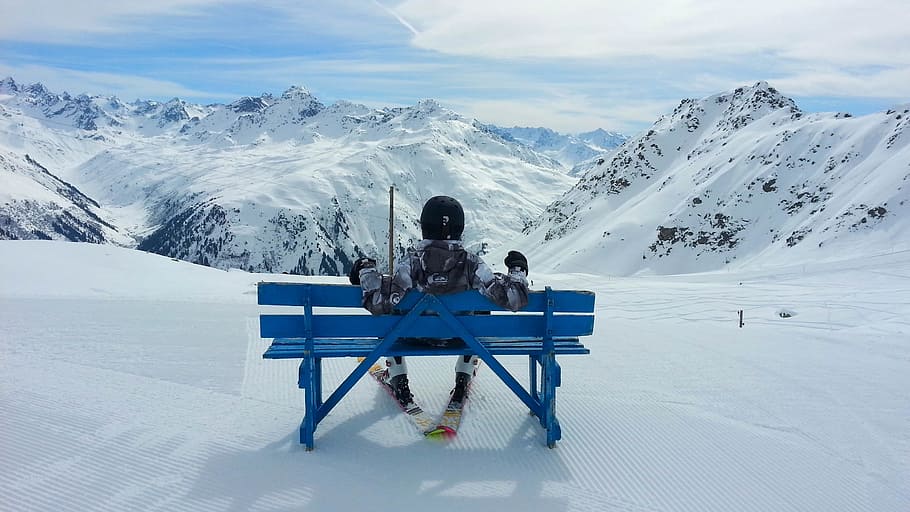 person sitting on blue wooden bench on ice coated place in front of mountains at daytime