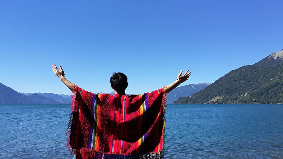 man wearing poncho with arms wide open in front of calm body of water and mountains during daytime