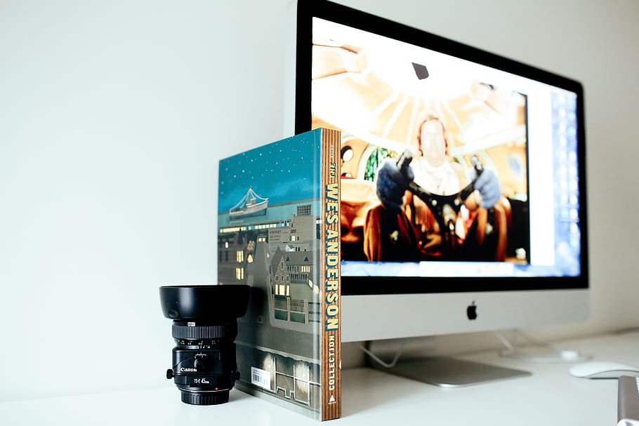 selective focus photography of black camera lens beside book and turned on iMac, silver iMac turned on beside book and camera lens