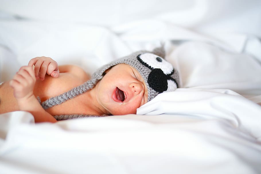 Yawning Baby, baby wearing black and white knitted owl critter hat lying on white textile