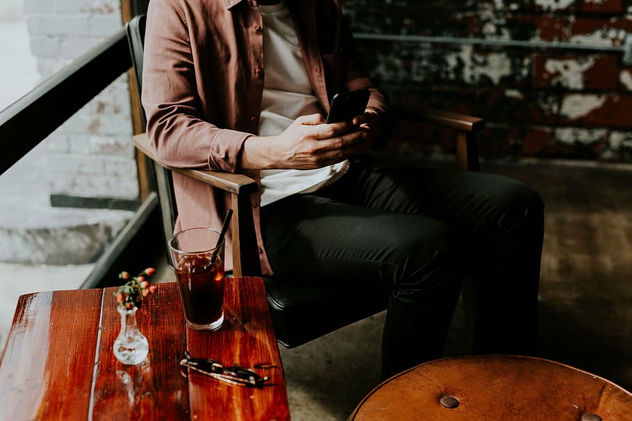 man holding black smartphone sitting on brown wooden armchair HD wallpaper