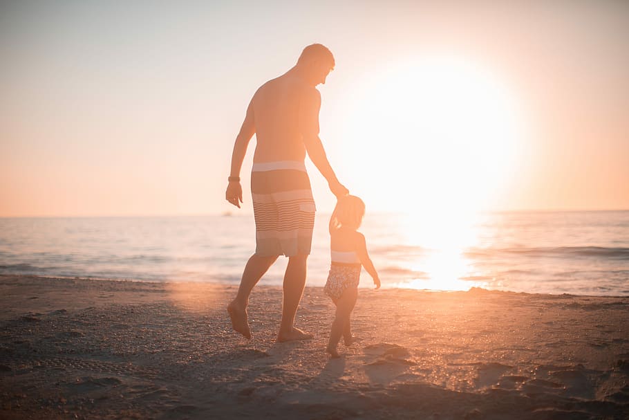 man holding girl heading towards sea, man wearing boardshorts holding toddler's left hand while walking on sand towards beach during sunset HD wallpaper