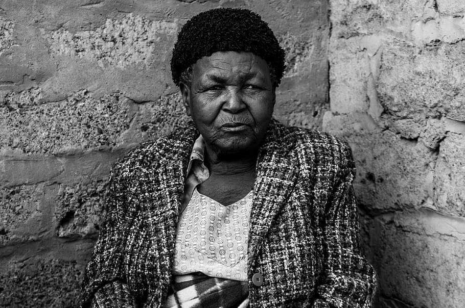Gogo Mabena, grayscale photo of woman wearing coat sitting beside concrete wall