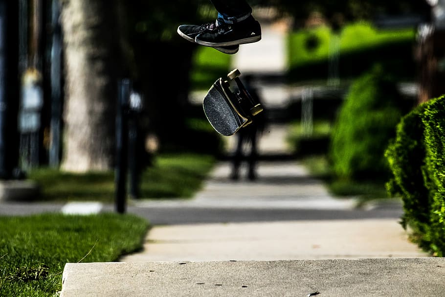black skateboard floating in mid air at daytime, person doing skateboard stunt in mid-air near green grasses and leafed plants during daytime