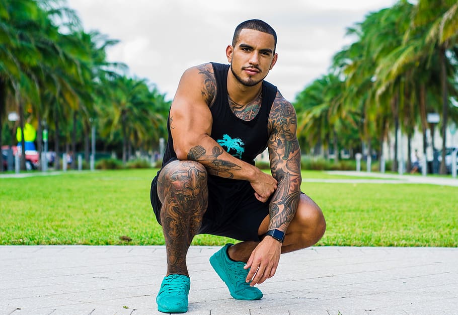 man in black tank top, black shorts, and pair of teal low-top sneakers, man in green grass field posing