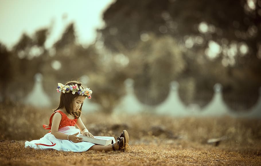 girl sitting whil holding book on field, girl wearing floral lei, red shirt, and white skirt sat on ground reading book