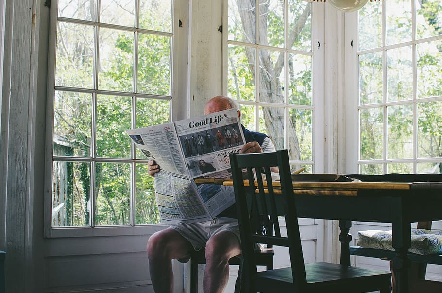 man reading newspaper while sitting on chair, man reading newspaper sitting beside table and glass window