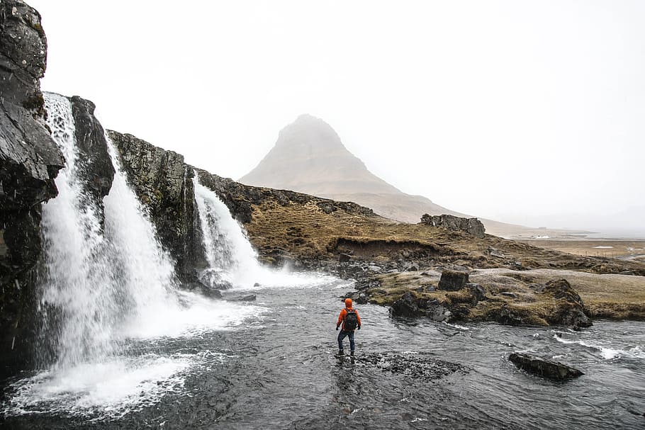 man with backpack standing on river beside waterfalls, photography of man in waterfalls outdoors HD wallpaper