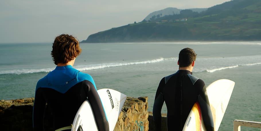two male wearing wetsuits holding surfboards standing nearby each other on top of hill with view of beach below, two man holding surfboards, HD wallpaper