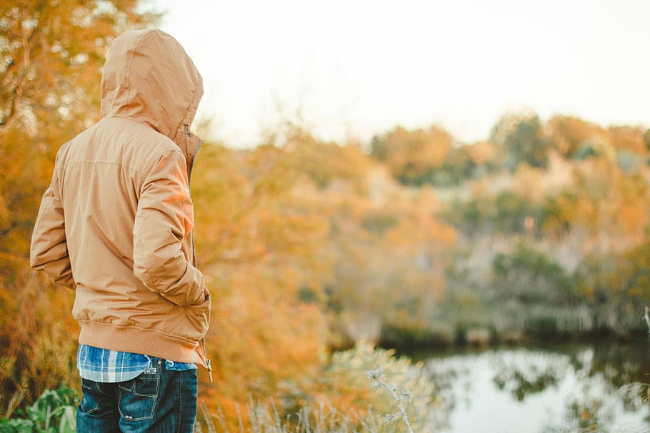 man in brown hooded jacket facing body of water during day HD wallpaper