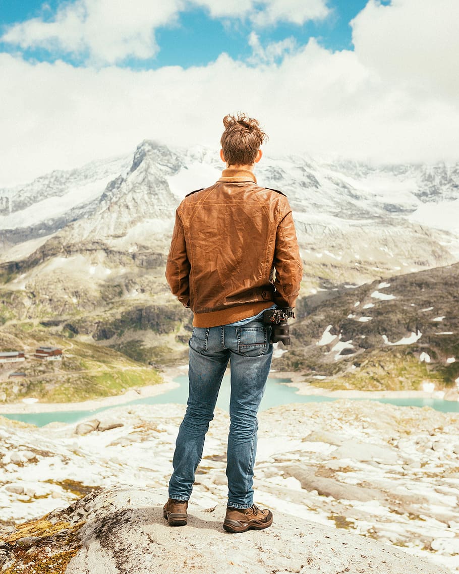 man standing on rock facing lake and mountains during daytime, man standing while watching the mountain HD wallpaper