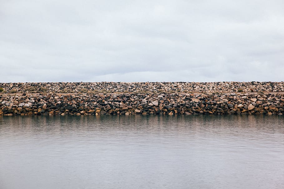 water rock wall during daytime, stack of rocks in front of body of water under white clouds HD wallpaper