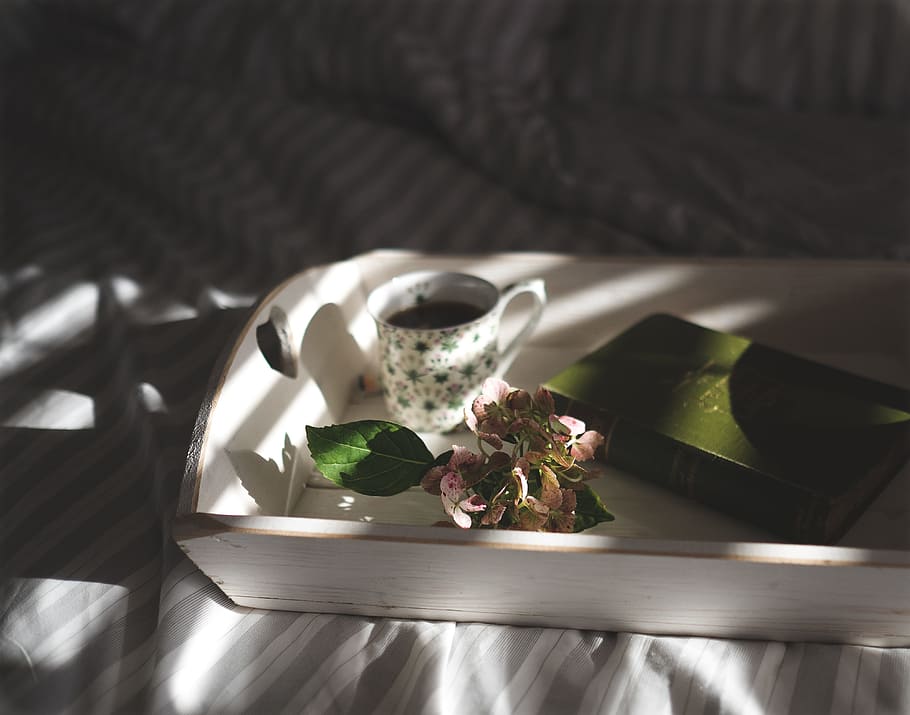 teacup beside pink flowers on tray, white wooden tray with white ceramic cup filled with coffee, green book, and bouquet of pink flowers on top