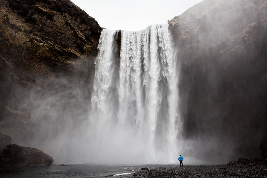 man standing front of waterfalls at daytime, brown rocky mountain and falls HD wallpaper