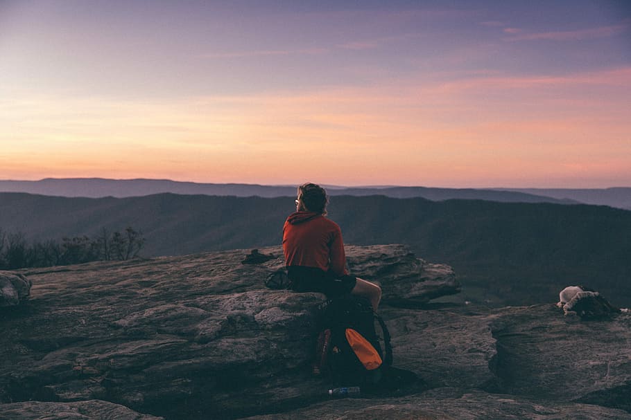 person sitting on boulder overlooking mountain during golden hour, person sitting on gray stone at sunset HD wallpaper
