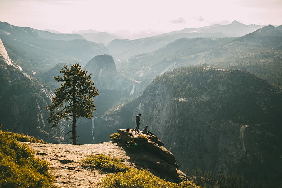 person standing on cliff, two persons sitting on edge of cliff during daytime HD wallpaper