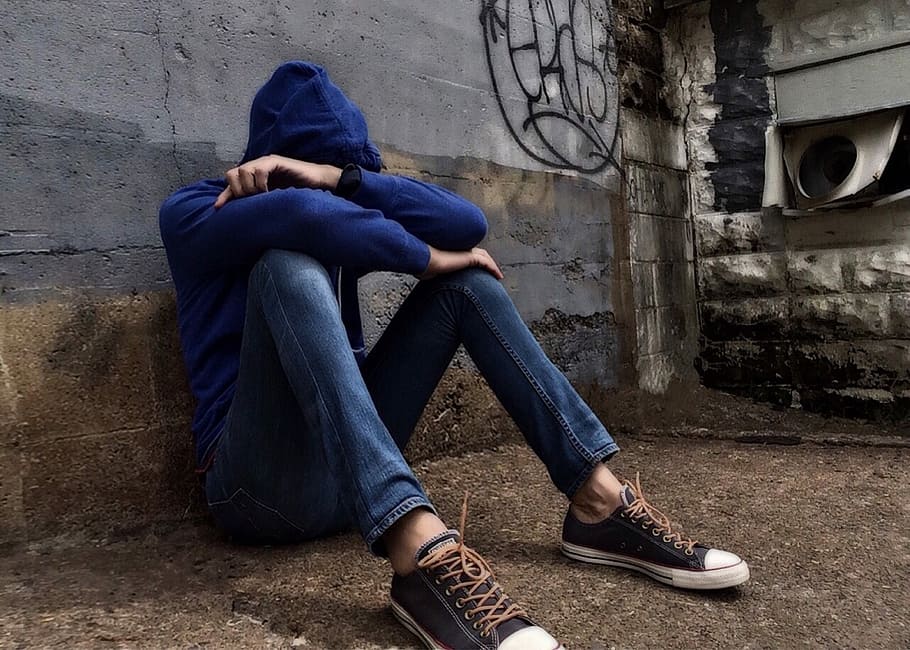 person in blue pullover hoodie and blue jeans sitting while leaning on gray concrete wall during daytime, HD wallpaper