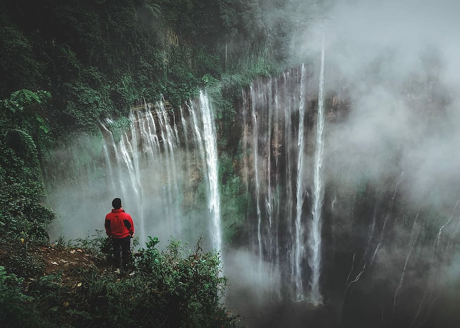man standing on cliff facing waterfall, person wearing red jacket standing near mountain cliff facing on waterfall at daytime HD wallpaper