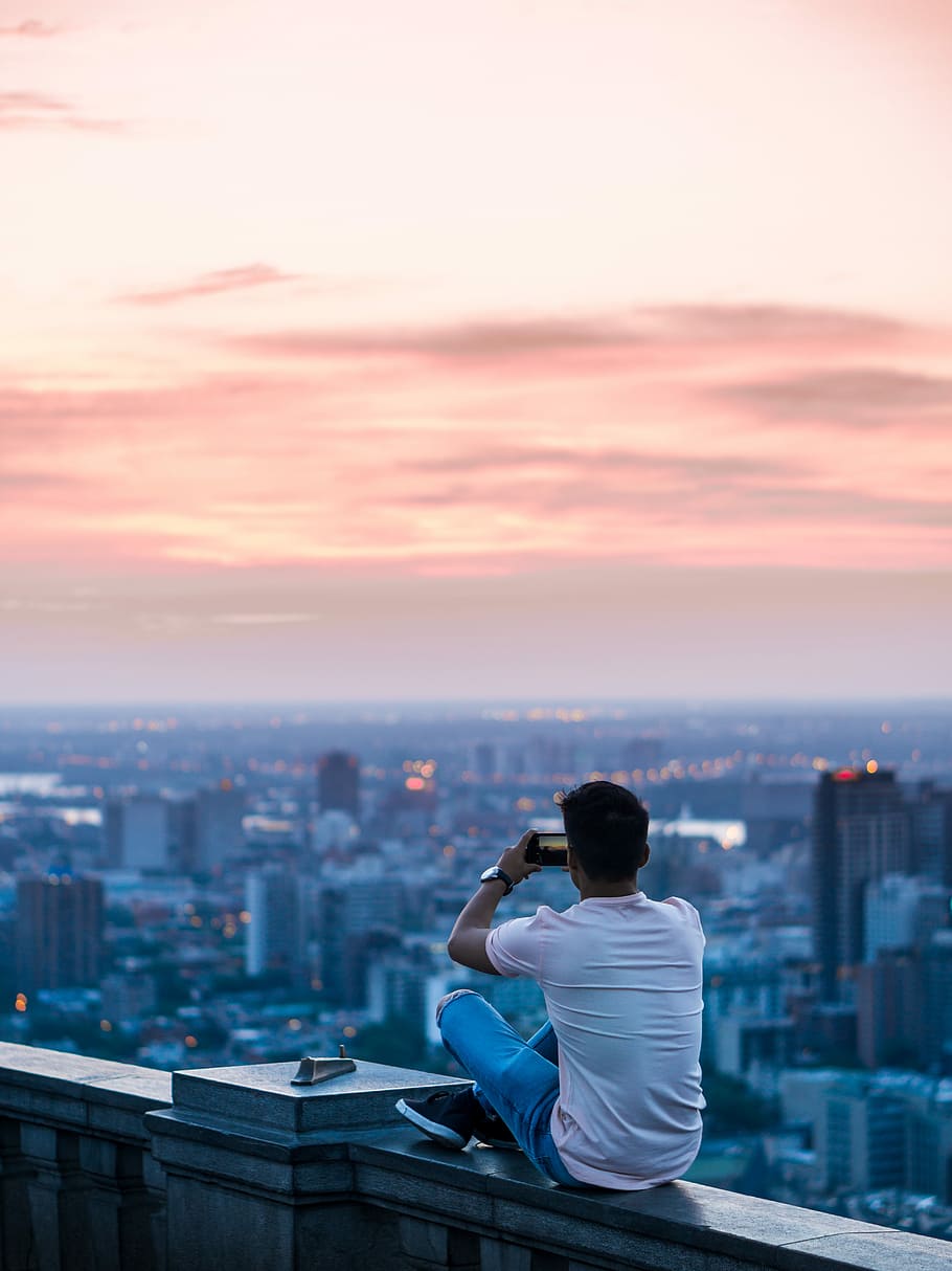 person sitting on edge of building taking photo of city during daytime, man sitting on gray concrete edge during sunset, HD wallpaper