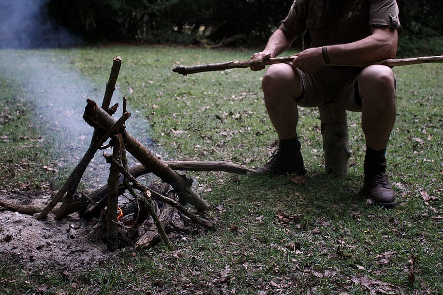 man holding brown stick, man sitting on log making fire with wood pile during daytime HD wallpaper