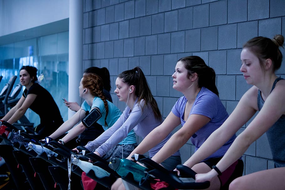 women taking exercise on black stationary bikes in front of gray concrete wall, people at gym room