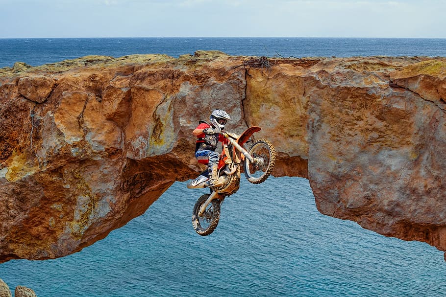 man riding motocross dirt bike in mid air near stone bridge and ocean during day