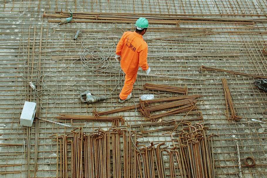 man walking on construction site, man in orange suit wearing teal hard helmet standing beside brown steel rods